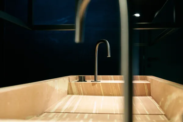 Waschbecken aus hellem Naturstein mit eleganter Maserung und modernem, gebogenem Wasserhahn – stilvolles Interior-Detail im ALEA Resort
