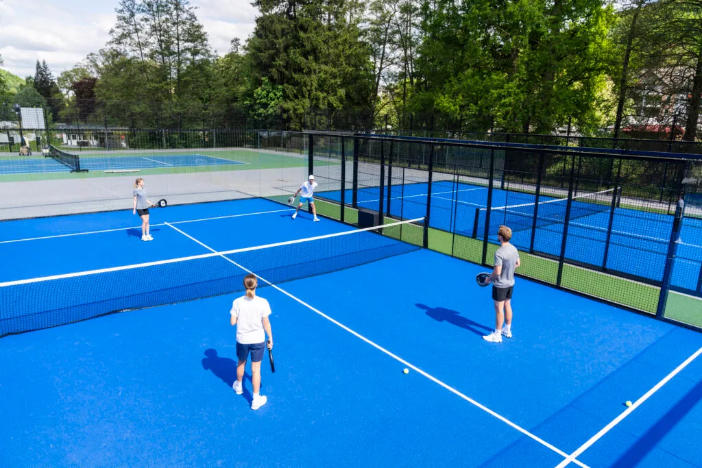 Vier Personen spielen ein Doppel-Match auf einem modernen blauen Padel-Tennisplatz im Freien, umgeben von Bäumen und Sportanlagen