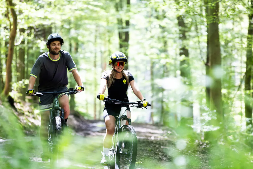 Zwei Personen fahren mit E-Mountainbikes durch einen sonnendurchfluteten Wald – aktive Erholung in der Natur